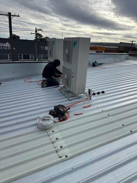 Condensing units being installed on the roof of a shop on a retail strip at dusk.
