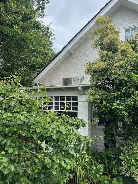 Outdoor condensing unit mounted high on brackets against a weatherboard wall.