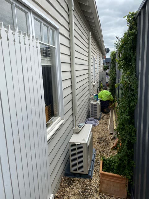 Two outdoor condensing units mounted side by side against a weatherboard wall.