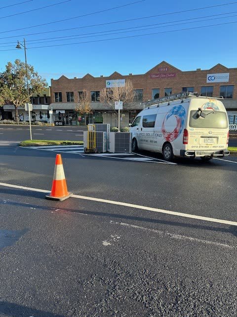 Branded Tailored Solutions van parked kerbside at a commercial job on a shopping strip, with outdoor plant and a traffic cone beside it.